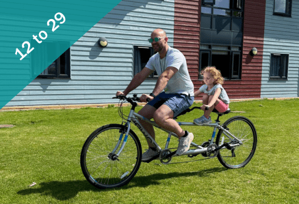 A father and daughter on a tandem bike on grass in sunshine. 12 to 29.