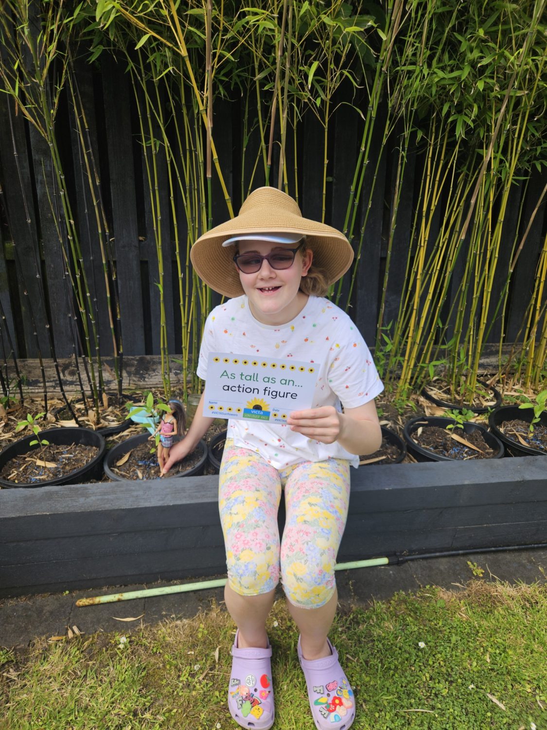 Felicity-action-figure Felicity sits on a raised planter, she wears a sun hat and sunglasses. She holds a sign ready 'as told as an action figure'. A Barbie sits next to the plant.