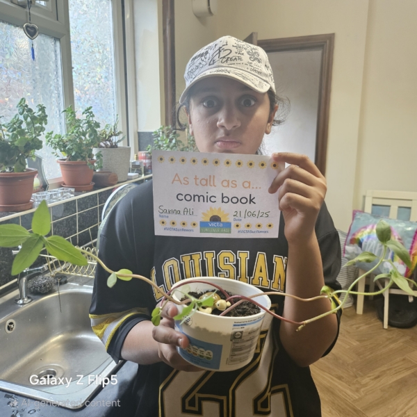 Sakina-comic-booked A teenager holds up a plant in her kitchen - it's growing tall outwards over the sides. She holds a sign reading 'as tall as a comic book.'