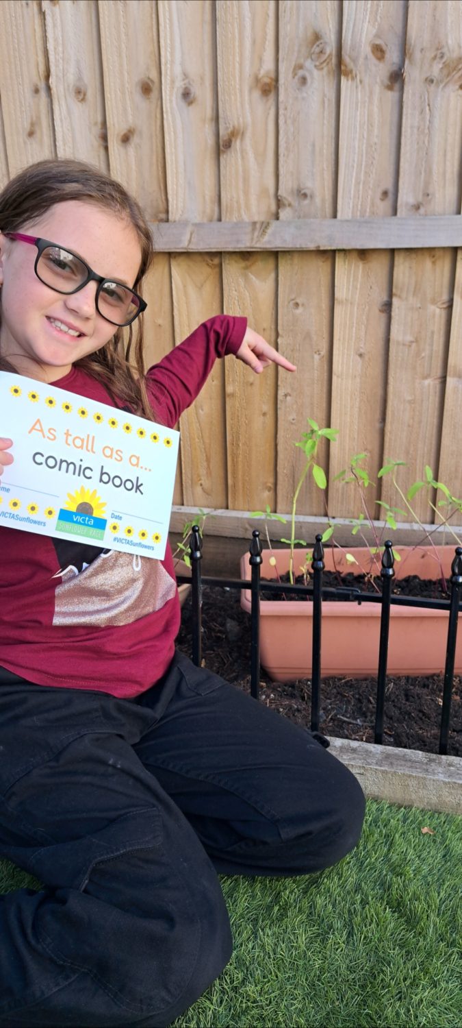 elena-comic-book Elena crouches down in her garden next to her sunflower plants. She points at the plant and holds up a sign reading 'as tall as a comic book'.