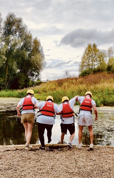 ellie-cowin The backs of Ellie and a group of volunteers, arm in arm about to step into the boating lake wearing their buoyancy jackets and helmets
