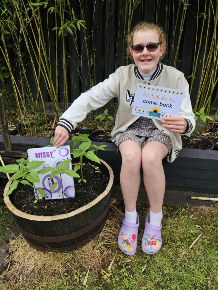 felicity-comic-book A teenage girl sits on a raised flower bed with a big smile. She holds her braille missy comic next to her sunflower plant and in the other hand a sign reading 'as tall as a comic'