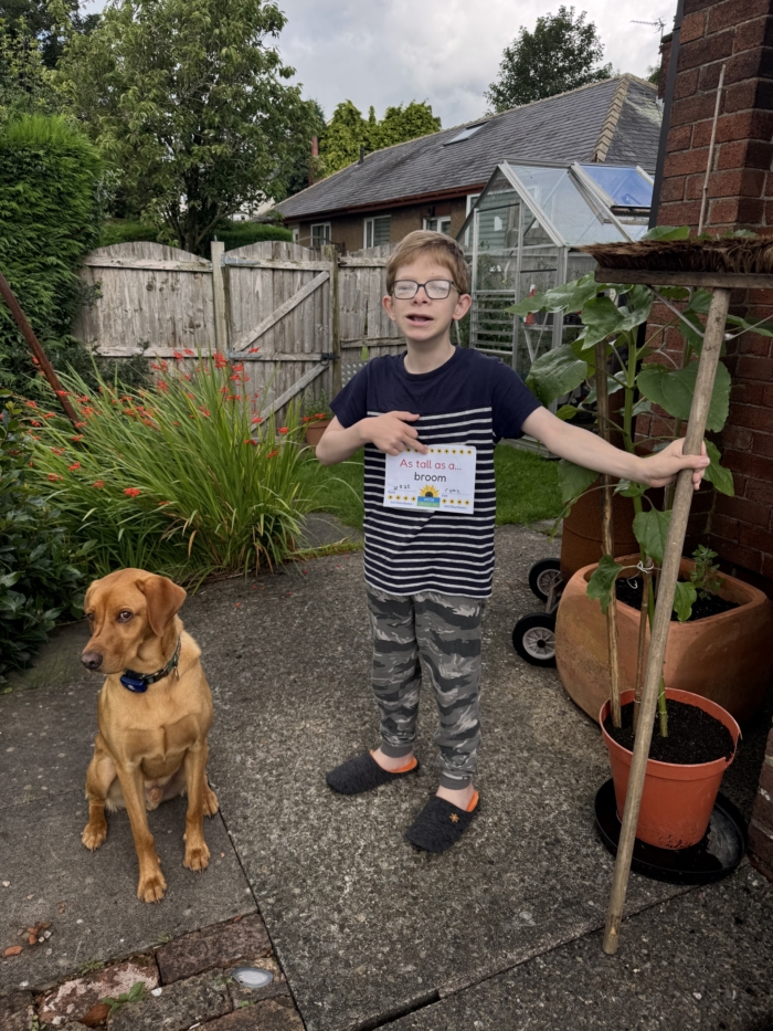 ryan-broom Boy holds a broom next to a sunflower plant with a sign reading 'as tall as a broom'. His dog sits obediently next to him.