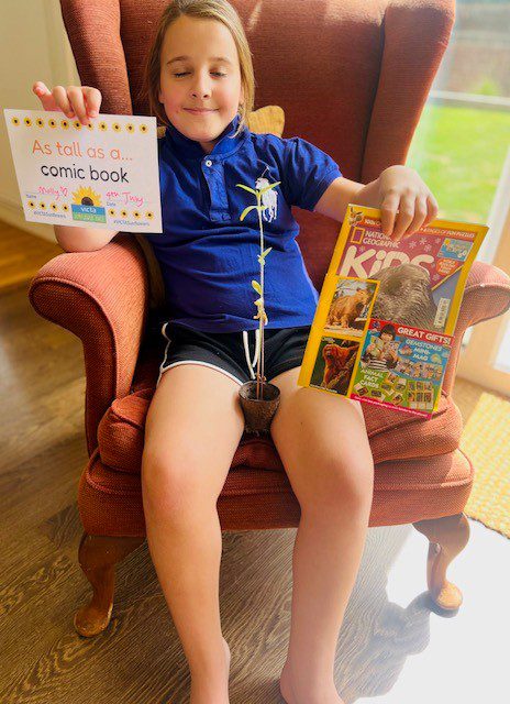 molly-2 Girl sits on an armchair with a sunflower plant on her lap. One hand holds a comic and the other a sign reading 'as tall as a comic'.
