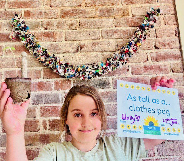 molly-3 Girl holds up a sunflower plant in one hand and a sign reading 'as tall as a clothes peg' in the other. She stands in front of an exposed brick wall with a pretty floral garland hanging from it.