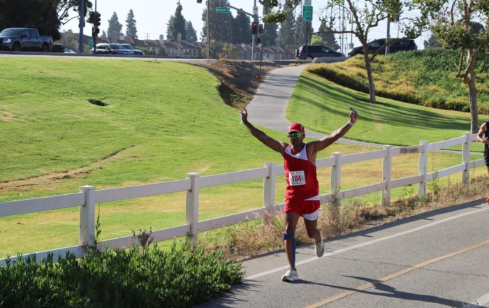 A runner in a red athletic outfit raises both arms in celebration while running on a sunny roadside path bordered by a white fence, with grassy hills and light traffic in the background.