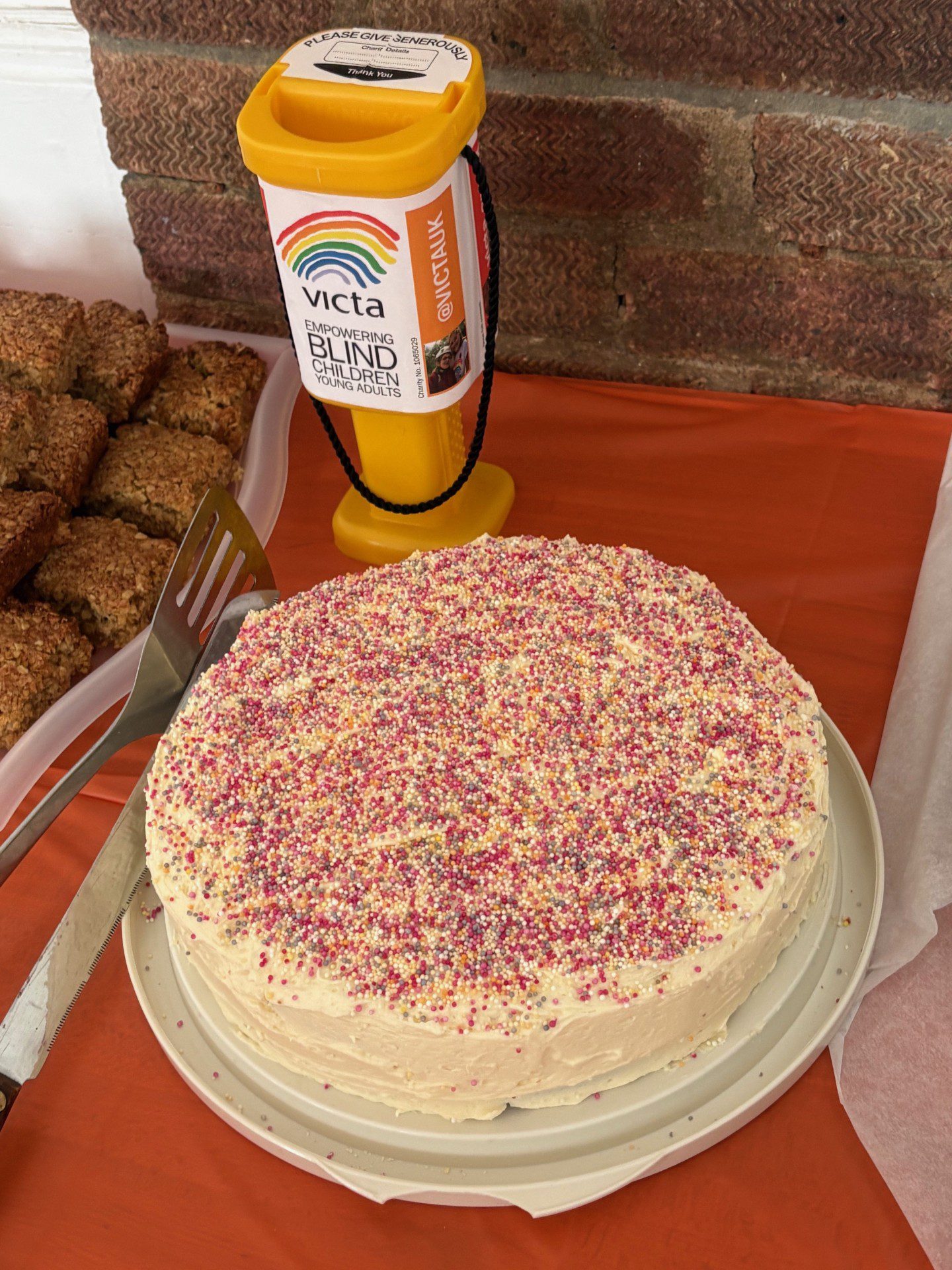 A round cake with white icing and rainbow sprinkles sits next to a VICTA donation goblet.