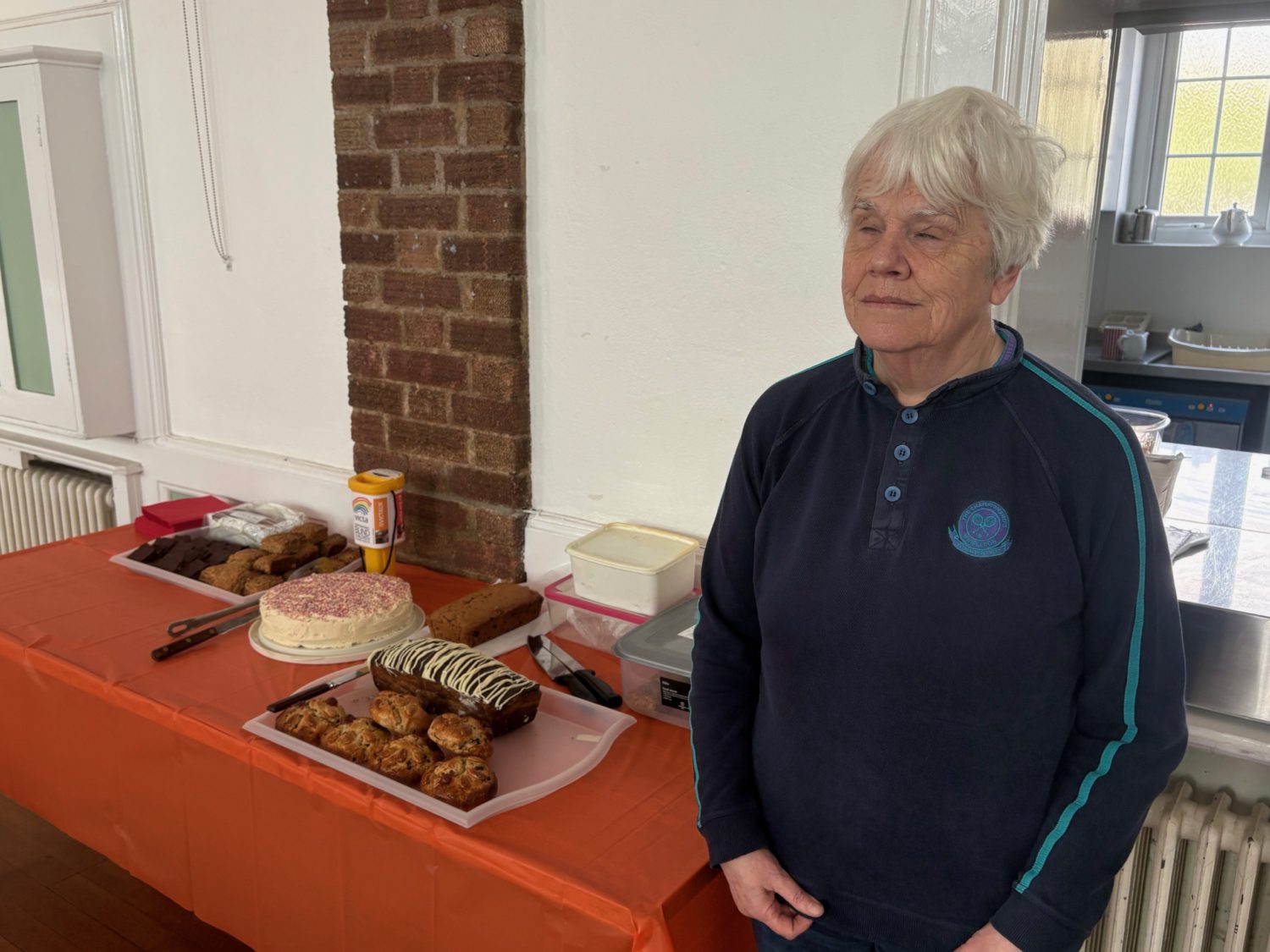 Mary smiles next to the cake table that has a selection of cakes and bakes!