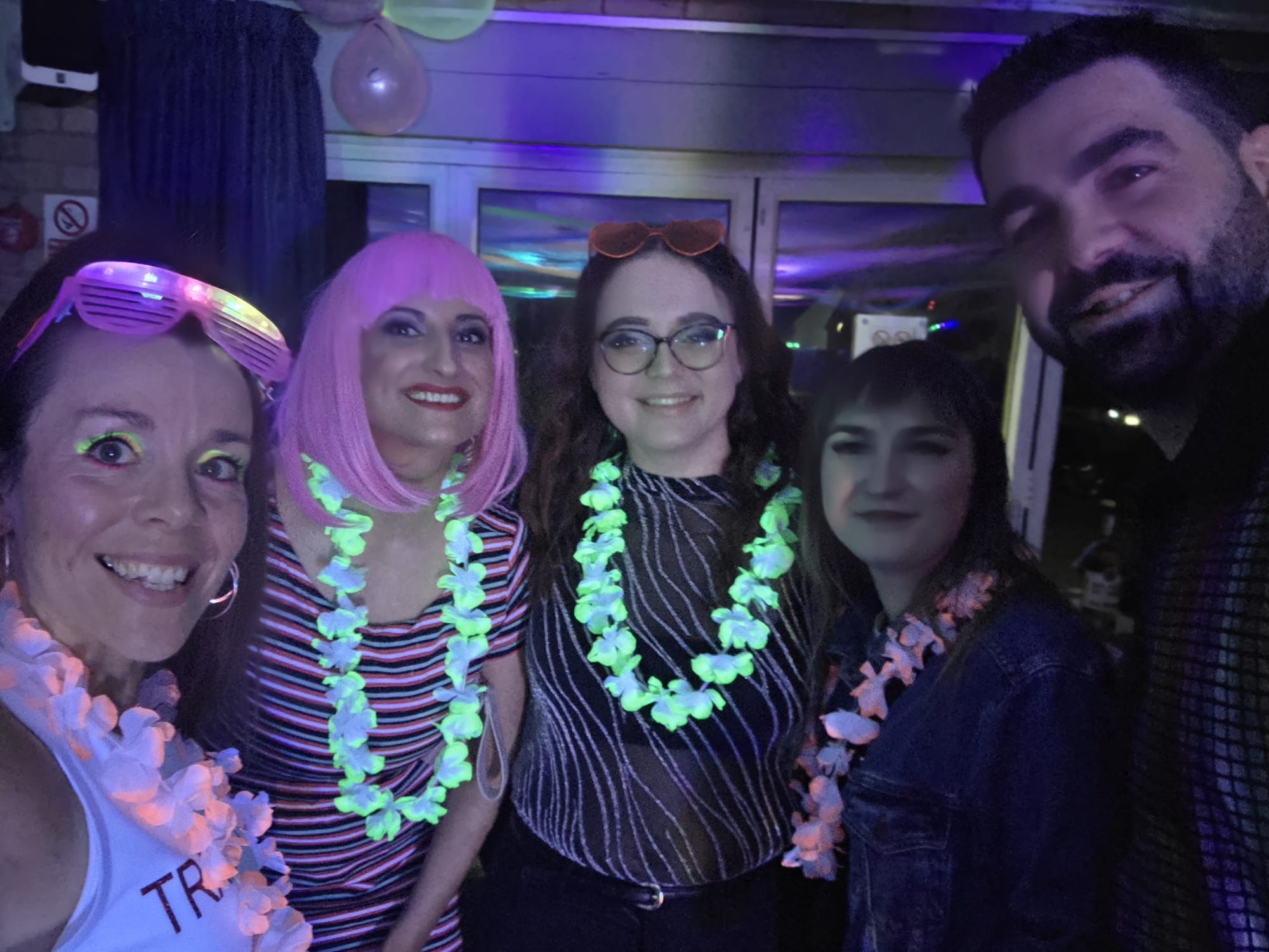 A group of friends smile in dark lighting wearing glowing UV flower garlands