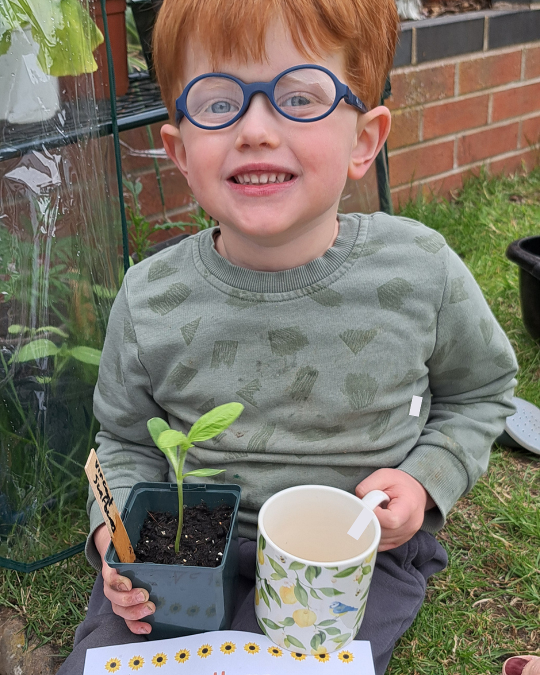 Boy smiling in a garden holding a flowerpot up next to a mug.