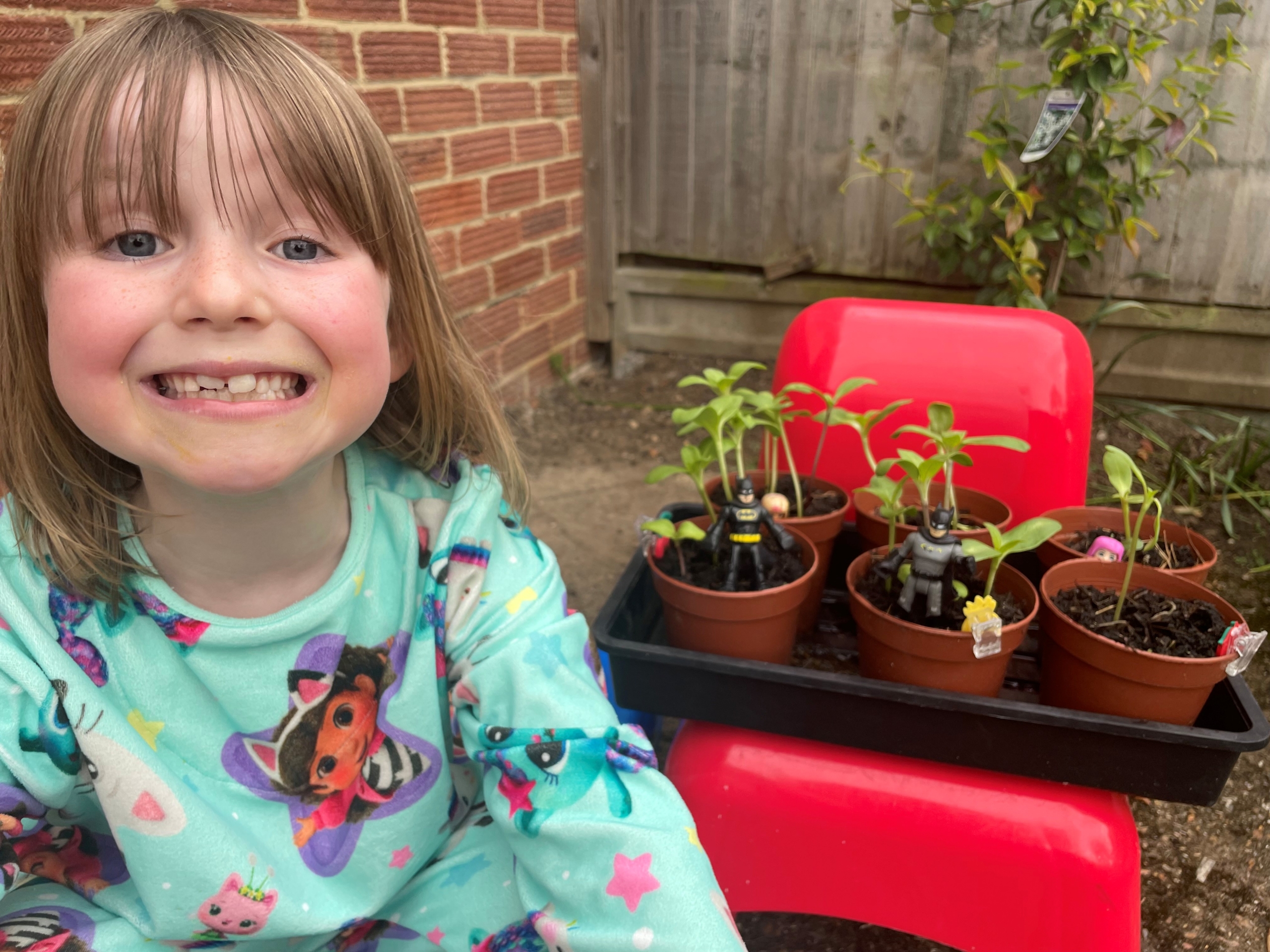 Girl smiling at the camera next to sunflower saplings as tall as mini figures in red pots.
