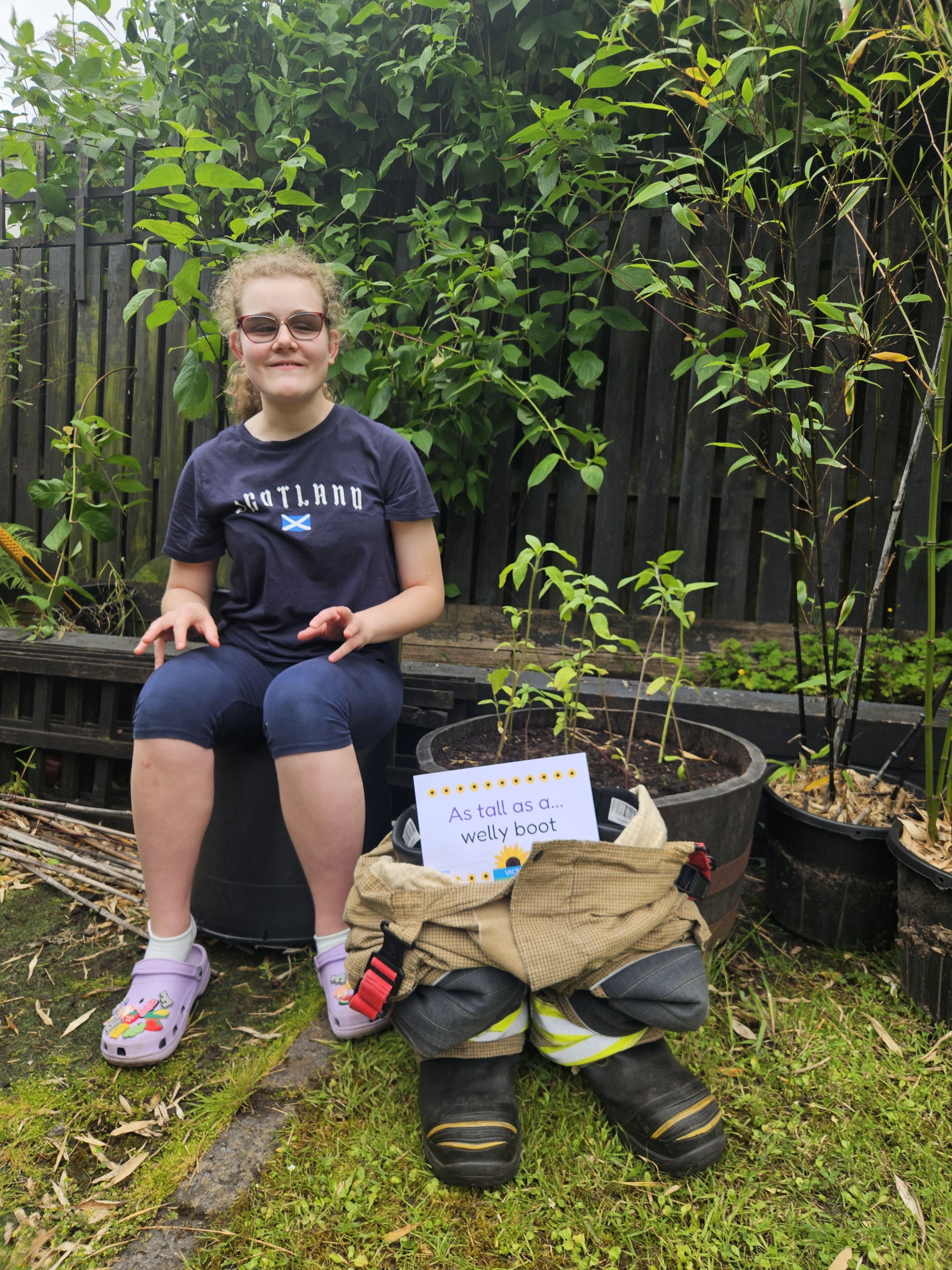 Felicity welly boot A girl sitting in a garden next to a sunflower growing from a pot as tall as a welly boot.