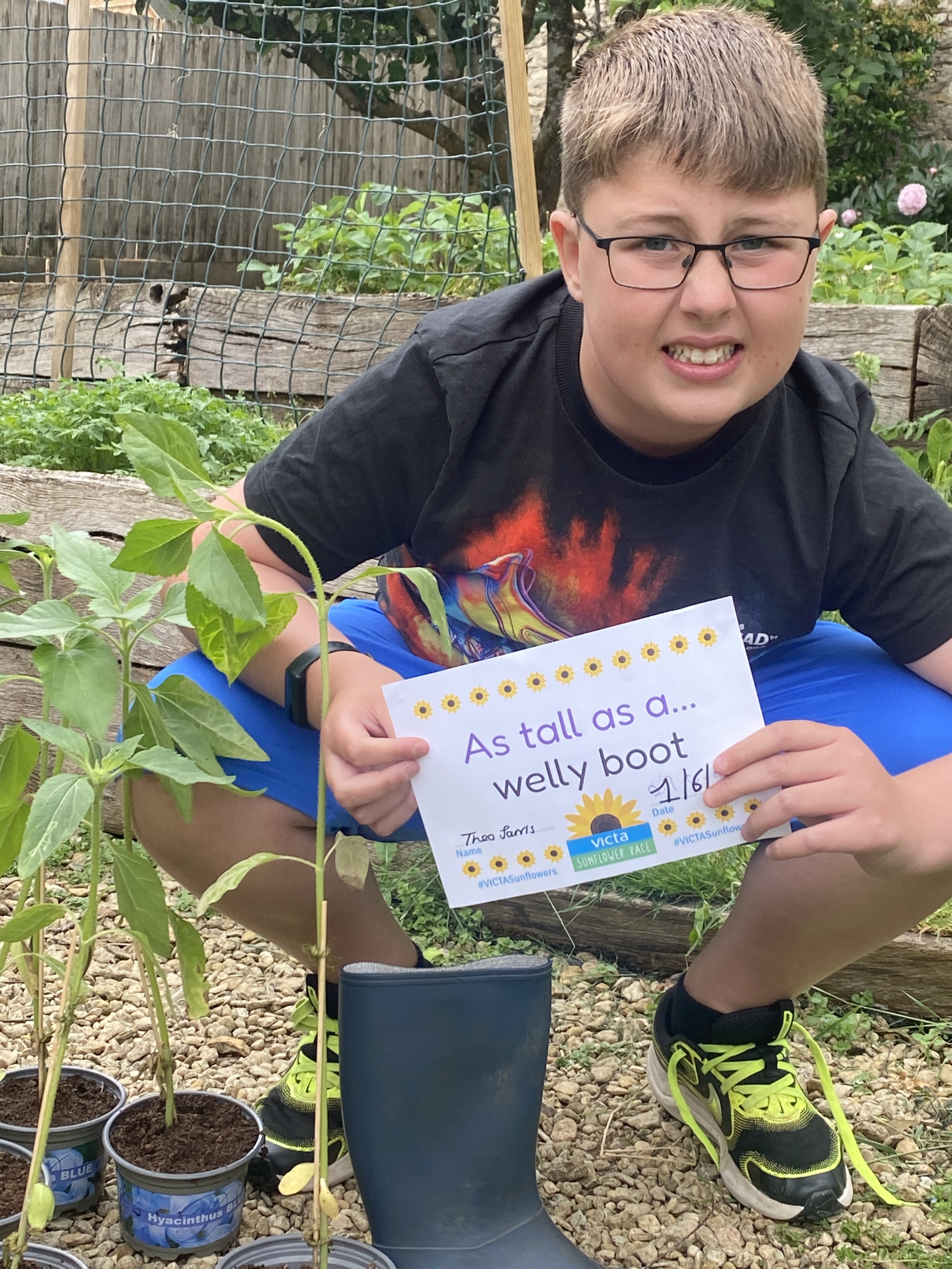Theo A boy wearing glasses and smiling crouched by a potted sunflower.