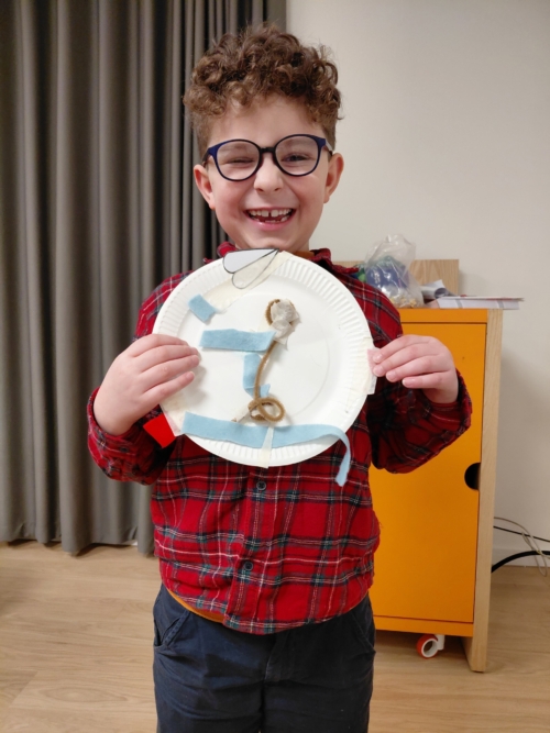 A boy has the biggest smile while he holds up a paper plate withe a tactile picture he has made.