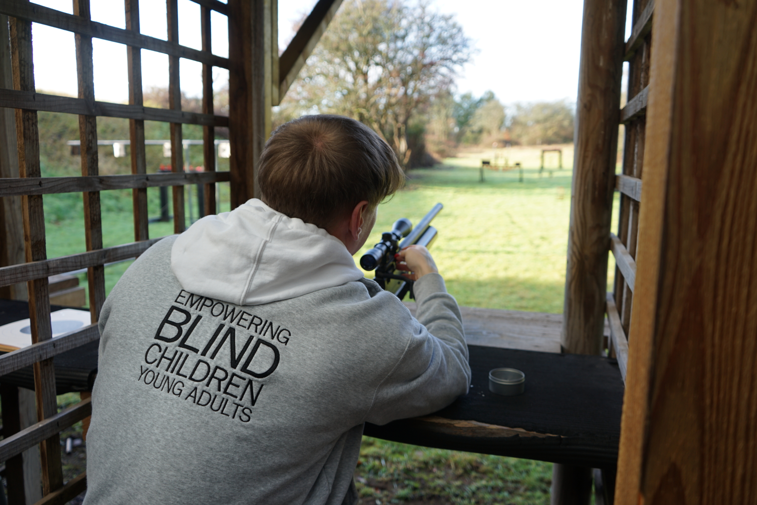 DSC00029 A male point there rifle through an outdoor shooting range. The have the words empowering blind and partially sighted on their back
