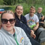 VICTA volunteers smile next to the raft building lake at summer camp